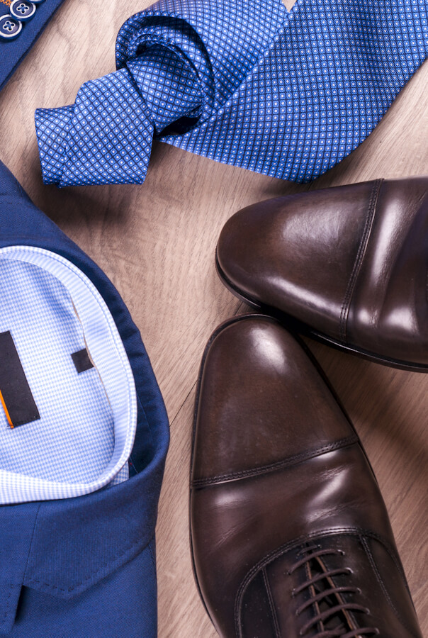 Flat lay set of classic mens clothes such as blue suit, shirts, brown shoes, belt and tie on wooden background